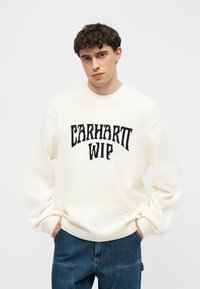 Jeune homme aux cheveux courts et bouclés portant un pull blanc Carhartt WIP et un jean bleu, mains dans les poches, sur un fond uni.