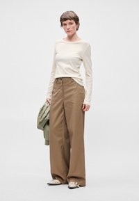 Κρέμα μπλούζα με μακριά μανίκια, καφέ παντελόνι πλατύς πόδι, πράσινο σακάκι ριγμένο στον ώμο και ανοιχτόχρωπα παπούτσια με λεπτομερείς λεπτομέρειες. Απλός, σύγχρονος σχεδιασμός.