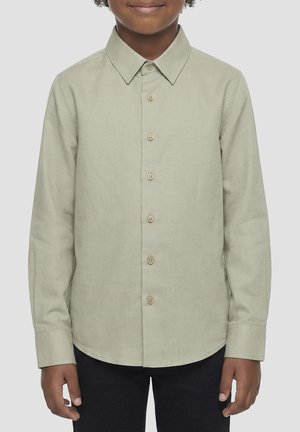 Niño con camisa de manga larga beige claro con botones y pantalones oscuros, de pie mirando hacia adelante con los brazos relajados a los lados.