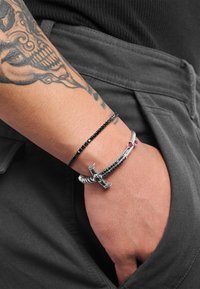 Schwarzes Perlenarmband und ein silbernes Armband mit roten Akzenten, verziert mit einem Kreuzanhänger, getragen an einem Handgelenk mit sichtbarem Tattoo.