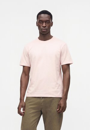 Ung mand med kort hår iført en lyserød kortærmet t-shirt og olivengrønne bukser, der står mod en ensfarvet hvid baggrund.