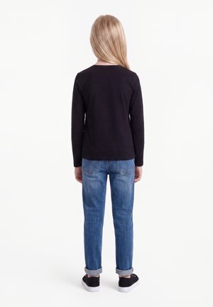Kind mit langen blonden Haaren steht mit dem Rücken zur Kamera, trägt ein schwarzes langärmeliges Shirt, blaue Jeans mit Umschlag und schwarze Sneaker mit weißen Sohlen.