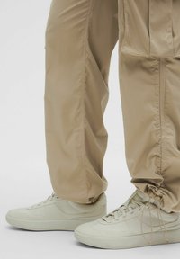 Beżowe, prążkowane spodnie z regulowanymi mankietami, połączone z jasnobeżowymi trampkami. Oba elementy mają gładką teksturę i minimalistyczny design.