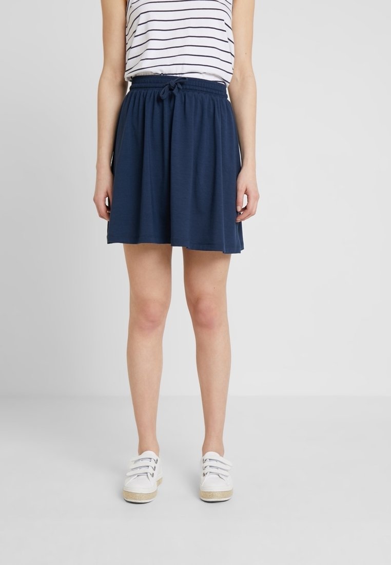 Femme portant une jupe bleu marine à cordon, un haut sans manches rayé blanc, et des baskets blanches, debout devant un fond gris clair uni.