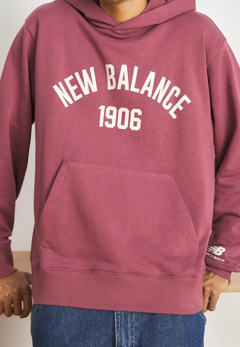 Personne portant un sweat à capuche New Balance bordeaux avec un texte blanc "NEW BALANCE 1906" et une poche avant, associée à un jean bleu.
