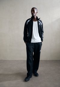 Black athletic jacket with gray accents, paired with a white tank top and black pants, worn with dark sneakers, set against a light background.