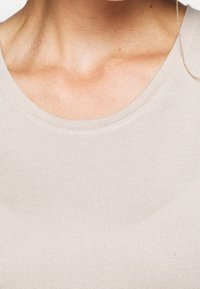 Světle béžová, ribkovaná látka s kulatým výstřihem a jemným řetízkovým detailem. Hladká textura, přiléhavý design, zaměřuje se na oblast límce.