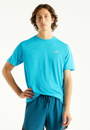 Jonge man die een lichtblauw sport-T-shirt en donkerblauwe korte broek draagt, staat met één hand op de heup tegen een witte achtergrond.