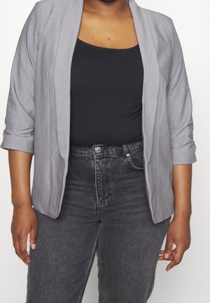 Person iført en grå blazer over en sort top og højttaljede mørkegrå jeans foran en ensfarvet hvid baggrund.