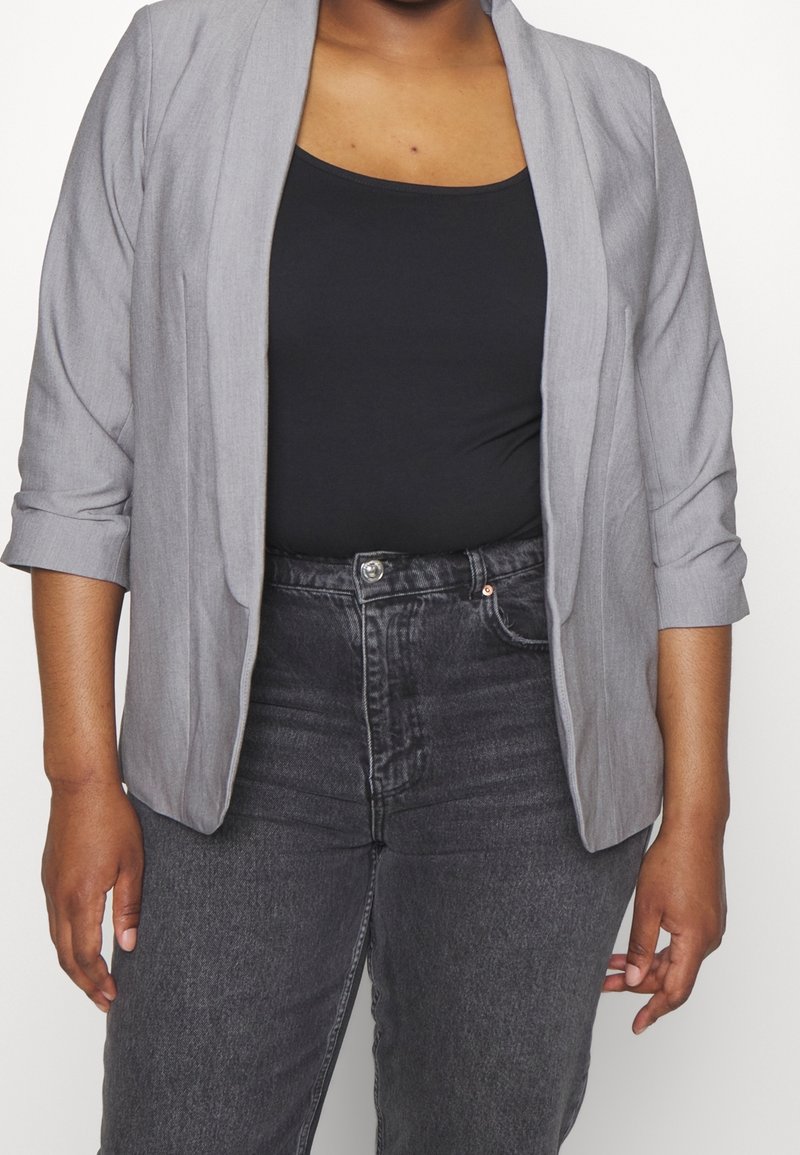 Personne portant un blazer gris sur un haut noir et un jean gris foncé taille haute sur un fond blanc uni.