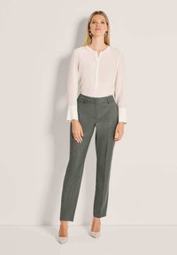 Vrouw die tegen een lichte muur staat, gekleed in een witte blouse met lange mouwen en brede manchet, grijze op maat gemaakte broek en beige puntige hakken.