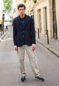 Blazer marine avec deux poches avant, porté sur un t-shirt noir, associé à un pantalon beige clair et des chaussures de sport noires, debout sur une rue de la ville.