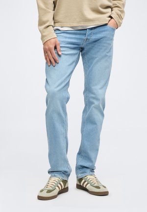 Mann trägt hellblaue Jeans, beigen Pullover und grüne sowie beige Sneaker, eine Hand in der Tasche, die andere auf dem Oberschenkel ruhend.