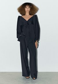 Zwarte langemouwen top met wijde halslijn en gerimpelde accenten, gecombineerd met een losse zwarte broek en platte zwarte sandalen.