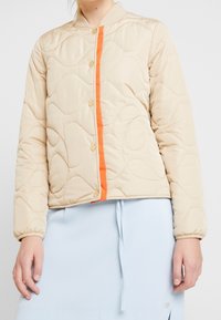 Beige prošívaná bunda s oranžovým pruhovaným motivem vpředu, zdobená zlatými knoflíky a žebrovaným límcem. Hladká textura, zkrácený střih.