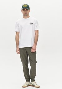T-shirt blanc à manches courtes avec un logo noir, pantalon vert olive, et une casquette verte avec un texte jaune. Les chaussures sont blanches avec des rayures noires.