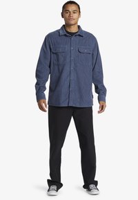 Camisa de pana azul con mangas largas, dos bolsillos frontales, cuello abotonado, combinada con pantalones negros y zapatillas negras.
