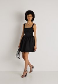 Melnas satīna mini kleita ar kvadrātveida dekoltē, pieguļošu korpusu un platu svārku. Modelis tur mazu sudraba rokassomu, valkājot melnas sandales ar siksniņām.