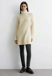 Kreminės spalvos virvės raštuotas megztinis su aukšta apykakle, derinamas su juodomis siaurėjančiomis kelnėmis ir juodais bateliais su raišteliais. Lygus paviršius, kasdieniško dizaino.