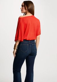 Rode gebreide top met een losse pasvorm en een off-shoulder ontwerp, gecombineerd met donkerblauwe slim-fit jeans met achterzakken en stikdetails.