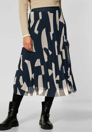 Plooirok in marineblauw met abstracte beige patronen. Heeft een brede tailleband en valt net onder de knie. Gedragen over zwarte leggings en laarzen.