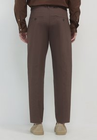 Mann trägt braune Anzughose und ein passendes Hemd mit beigen Slippern, stehend vor einem schlichten hellen Hintergrund.
