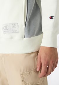 Felpa leggera color crema con accenti grigi, caratterizzata da un toppa con logo e polsini a costine. Tessuto morbido e design sportivo evidenziato da cuciture laterali.