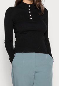 Femme portant un haut noir cintré à manches longues avec cinq boutons décoratifs et un pantalon taille haute bleu clair, les mains dans les poches.