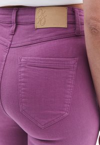 Jeans de mezclilla morados ajustados con un parche de cuero que presenta un logotipo de la marca en relieve en la cintura y un solo bolsillo trasero con detalles de costura.