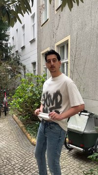 Homme portant un t-shirt graphique blanc et un jean bleu, debout sur un chemin pavé à proximité de buissons et d'une remorque pour vélo dans une cour urbaine.