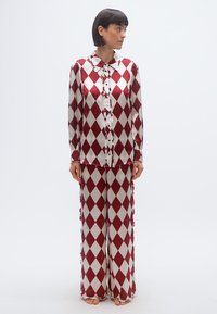 Femme debout pieds nus, portant un chemisier à manches longues et un pantalon large assortis, avec un motif de losanges rouges et blancs, regardant vers sa gauche.