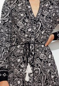 Robe avec motif paisley noir et blanc, à boutons sur le devant, taille nouée et accents à franges sur le cordon de serrage. Manches longues avec détail floral.