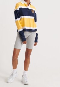 Maglietta da rugby a maniche lunghe con righe orizzontali blu navy, giallo e bianco, caratterizzata da un colletto bianco e un logo ricamato. Indossata con pantaloni corti grigi e sneaker.