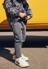 Kind in einer blumigen Steppjacke mit Reißverschluss, grauen Sweatpants und weißen Sneakern mit roten Akzenten vor einem gelben Hintergrund.