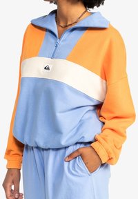 Pullover color block in arancione, azzurro chiaro e bianco. Presenta un colletto con zip, maniche lunghe e una vestibilità rilassata con polsini e tasca laterale.