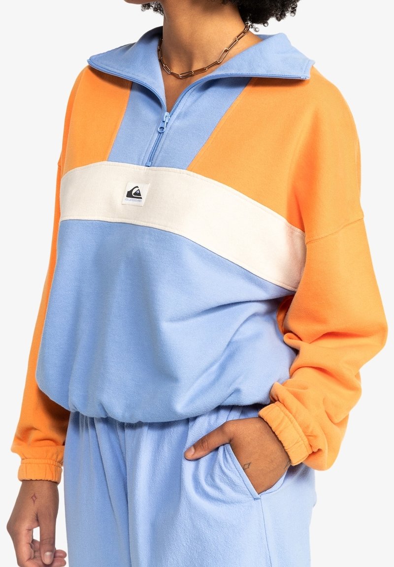 Pullover color block in arancione, azzurro chiaro e bianco. Presenta un colletto con zip, maniche lunghe e una vestibilità rilassata con polsini e tasca laterale.