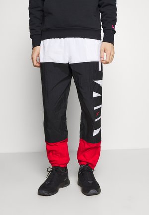 Mężczyzna ubrany w czarne, białe i czerwone dresy Nike w bloki kolorów oraz czarne sneakersy, stojący na jednolitym tle.
