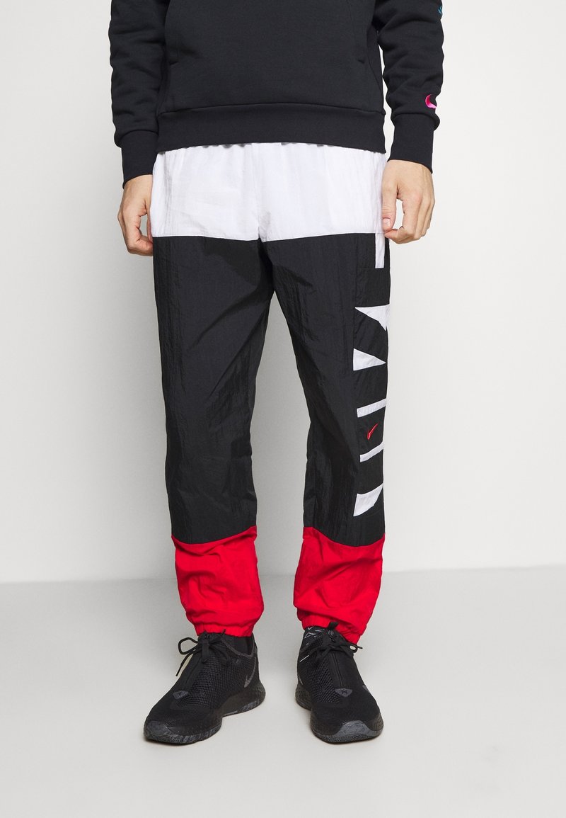 Mann in schwarz-weißen und roten color-blocked Nike Jogginghosen mit schwarzen Sneakers, der vor einem einfarbigen Hintergrund steht.