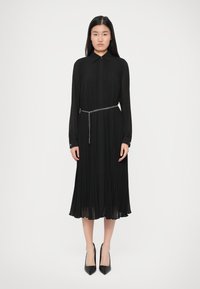 Γυναίκα που φοράει ένα μαύρο πλισέ midi φόρεμα με μακριά μανίκια και μεταλλική ζώνη αλυσίδα, καθώς και μαύρες ψηλές γυναικείες γόβες με μυτερή μύτη, στέκεται μπροστά σε απλό φόντο.