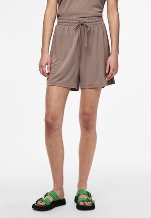 Persoon die taupekleurige shorts met trekkoord en groene sandalen met dubbele gesp draagt, staand tegen een egale witte achtergrond.