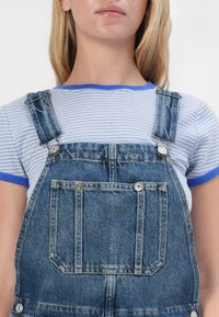 Jeanslatzhosen mit silbernem Zubehör, verstellbaren Trägern und einer Vordertasche, getragen über einem blau-weißen gestreiften T-Shirt mit kurzen Ärmeln.