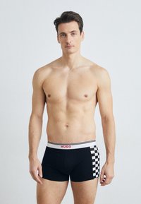 Boxeri negri cu un model pătrat pe o parte și un elastic alb cu "HUGO" în litere roșii. Textură de țesătură netedă.