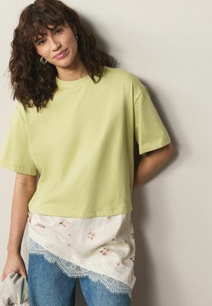 Mujer joven con cabello rizado que lleva una camiseta verde claro de gran tamaño sobre una blusa con encaje floral y jeans azules, sosteniendo un objeto doblado en la mano.