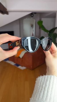 Sorte katteøje-solbriller med guld "DG"-logo holdt af to hænder i et hyggeligt rum med en brun sofa og en grøn plante i baggrunden.