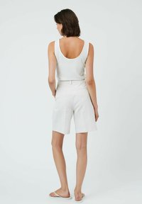 Weißes geripptes Tanktop mit einem runden Rückenausschnitt, kombiniert mit hellbeigen Leinen-Shorts. Modell trägt einfache flache Sandalen. Schlichter Hintergrund.