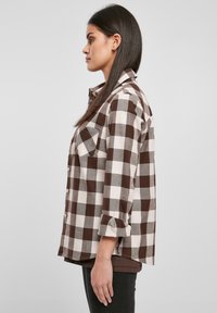 Camicia a quadri marrone e bianca con maniche lunghe, due tasche sul petto e chiusura con bottoni. Il tessuto sembra morbido con una finitura leggermente strutturata.