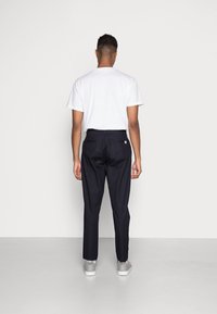 Mörk marinblå byxor med fram- och bakfickor, bärs med en enkel vit t-shirt och grå skor, sedd bakifrån.