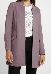 Femme portant un blazer ouvert texturé mauve sur un t-shirt blanc avec un imprimé abstrait de visage et un pantalon noir, debout devant un fond blanc.