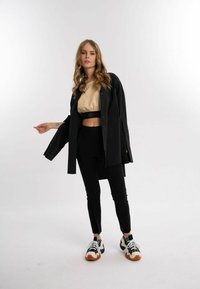 Sort skjorte med knapper over en beige cropped top, parret med sorte leggings og chunky sort-hvide sneakers. Glat tekstur og tætsiddende design.