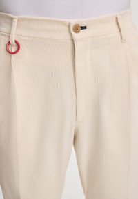 Beżowe sztruksowe spodnie o prążkowanej teksturze, z zapięciem na drewniany guzik i czerwoną metalową ozdobną obręczą na pasku.